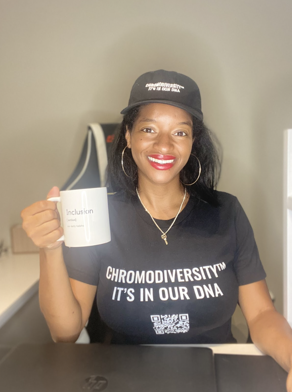 Jourdan is wearing red lipstick sitting in a black and white chair at her desk smiling, wearing a black shirt and hat with white text “Chemodiversity™ It’s In Our DNA.” On the shirt there is a QR code under the text. She is wearing a gold key necklace and holding a white mug with text “Inclusion action our daily habits.”